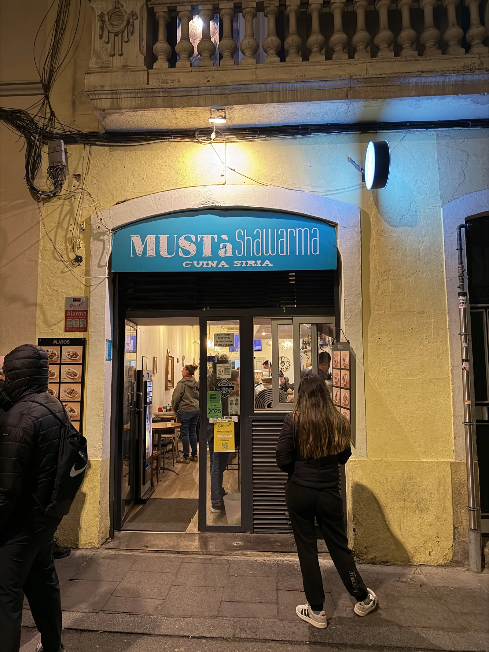 L'entrada de Mustà Shawarma al carrer de Mozart, de nit, amb el rètol blau